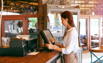 A hospitality worker stands at the counter of a cafe, typing into a computer.